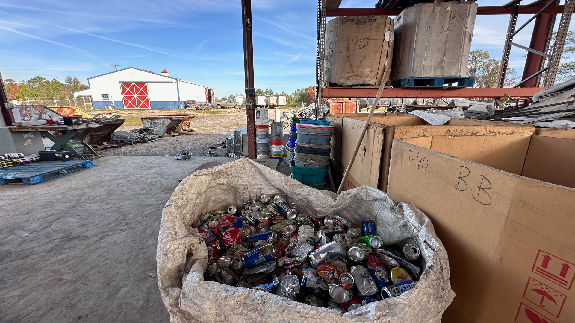Bulk bag of loose aluminum beverage cans in the covered processing shed beside labeled Gaylord boxes of lead and sorted small non-ferrous scrap, with the red-and-white CW barn office in the background